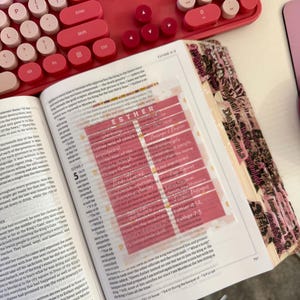 May include: Open Bible with a pink and white keyboard in the background. A clear, pink-tinted page marker highlights text. The Bible is open to the book of Esther, with decorative pink tabs.