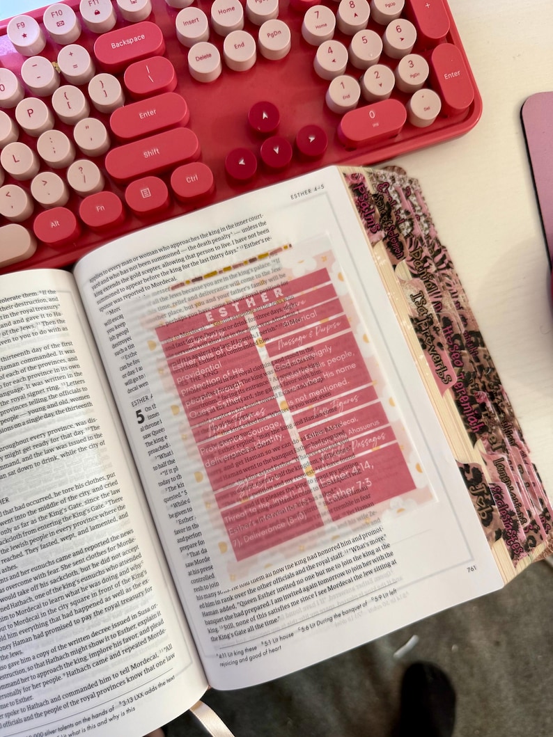 May include: Open Bible with a pink and white keyboard in the background. A clear, pink-tinted page marker highlights text. The Bible is open to the book of Esther, with decorative pink tabs.