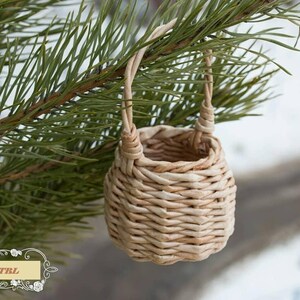 May include: A small, woven basket with a handle hangs from a pine tree branch. The basket is made of natural materials and has a light brown color.