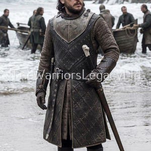 Puede incluir: Una persona con una túnica y armadura de cuero acolchado negro y marrón, sosteniendo una espada. El atuendo incluye una placa de pecho de metal y botas de cuero. La escena está ambientada en una playa con botes al fondo.