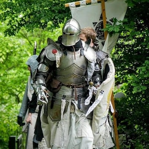 May include: A group of people in medieval armor, including helmets, chest plates, and leg protection, walking through a wooded area. One knight carries a banner with a black symbol. The armor is silver and the clothing is white.