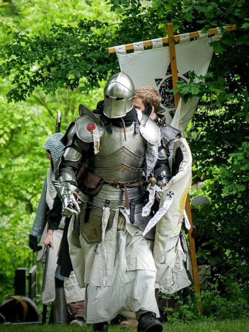 May include: A group of people in medieval armor, including helmets, chest plates, and leg protection, walking through a wooded area. One knight carries a banner with a black symbol. The armor is silver and the clothing is white.