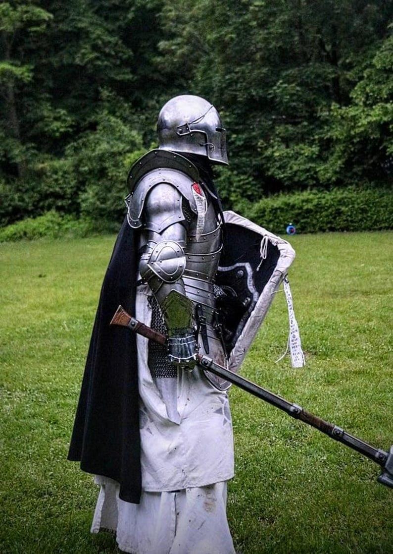May include: A person in full medieval armor, including a helmet, chest plate, and shoulder guards. The armor is silver-colored and worn with a black cape and white tunic. A shield and a polearm are also visible.