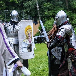 May include: Two people in medieval armor face each other on a grassy field. One holds a shield with a painted woman, the other a sword. The armor is silver, and the shield has a yellow halo.