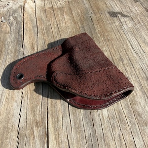 May include: A dark burgundy leather holster with a textured suede-like exterior. The holster has a curved shape with a stitched seam and a small hole for a belt loop. The interior is a smooth, darker shade of burgundy.