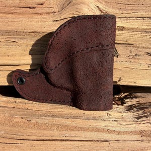 May include: A dark brown leather holster with a textured surface. The holster is stitched along the edges and features a small, circular metal rivet. The item is displayed on a wooden surface.