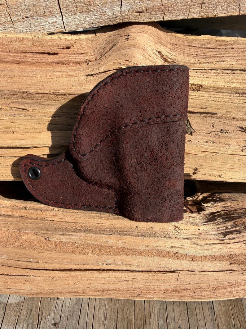 May include: A dark brown leather holster with a textured surface. The holster is stitched along the edges and features a small, circular metal rivet. The item is displayed on a wooden surface.