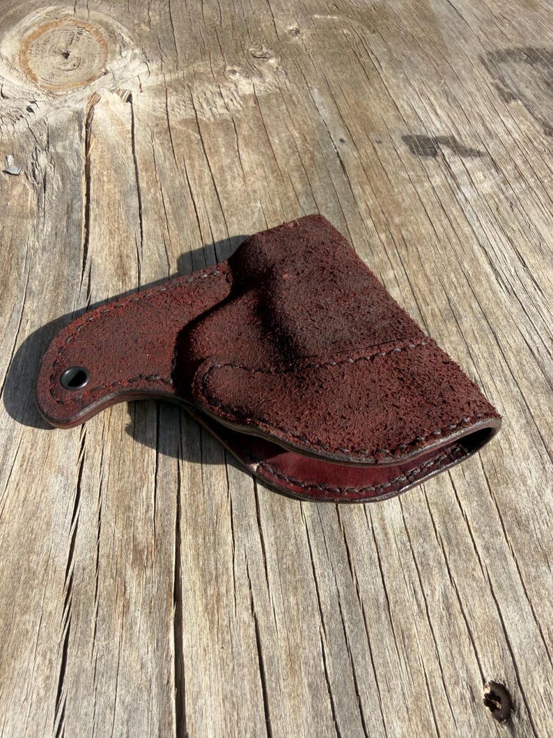 May include: A dark burgundy leather holster with a textured suede-like exterior. The holster has a curved shape with a stitched seam and a small hole for a belt loop. The interior is a smooth, darker shade of burgundy.