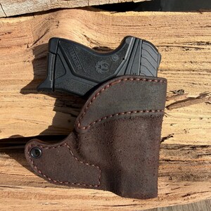 May include: A black handgun sits in a brown leather holster. The holster is stitched with red thread and has a dark brown suede texture. The handgun is a Ruger model.