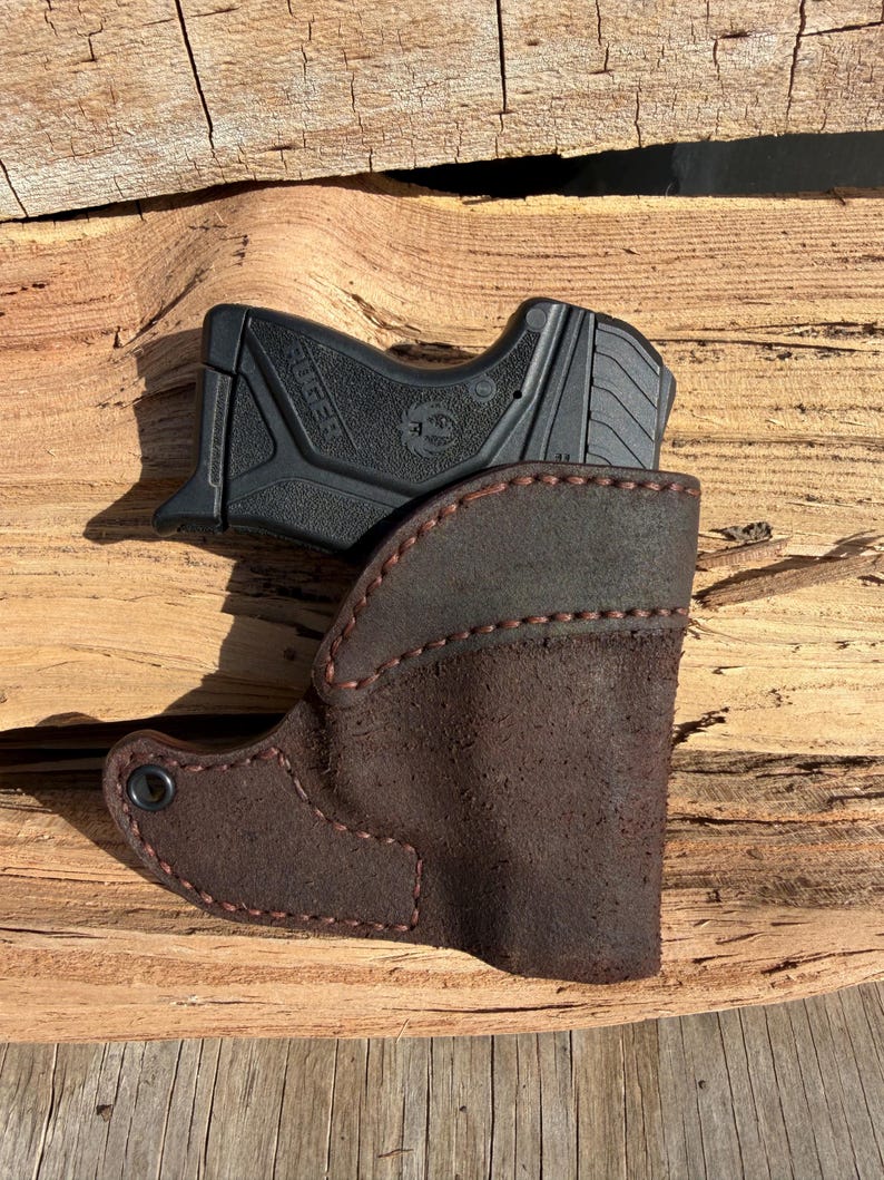 May include: A black handgun sits in a brown leather holster. The holster is stitched with red thread and has a dark brown suede texture. The handgun is a Ruger model.