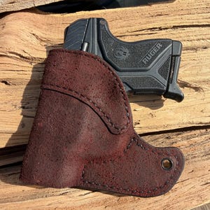 May include: A black Ruger handgun rests partially inside a burgundy leather holster. The holster is stitched with black thread and has a metal grommet. The handgun is black with the word "Ruger" printed on the side.