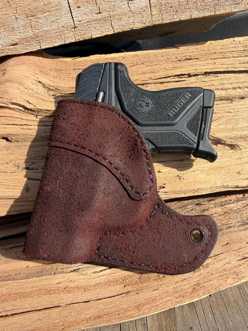May include: A black Ruger handgun rests partially inside a burgundy leather holster. The holster is stitched with black thread and has a metal grommet. The handgun is black with the word "Ruger" printed on the side.