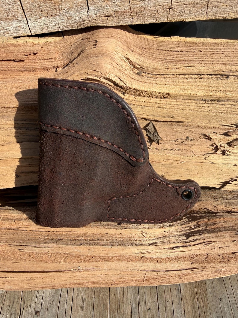 May include: A dark brown leather boot-shaped item with visible stitching. The boot has a suede-like texture on the main body and a smoother leather top. The stitching is a contrasting reddish-brown colour.