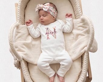 May include: A sleeping infant wearing a white long-sleeve bodysuit with a floral letter N and the name Nicole printed on the chest, paired with a matching floral headband, resting inside a cream-colored woven bassinet with a soft fabric liner.