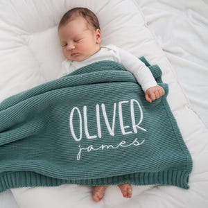 May include: A sleeping baby is nestled under a teal knitted blanket. The blanket is embroidered with the name "OLIVER" above "james" in white script. The baby is wearing a white onesie and resting on a white surface.