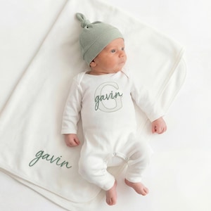 May include: A newborn baby wearing a sage green knotted hat and a white onesie with the name "Gavin" embroidered in green. A matching white blanket with the name "Gavin" is also present. The items are on a white background.