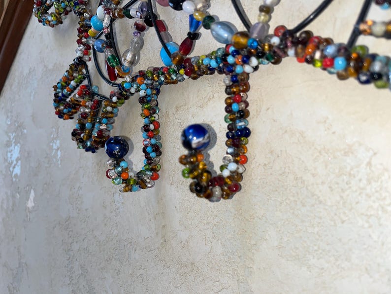 May include: Close-up of a decorative wall hanging with a black metal frame. The design features intricate loops and curves adorned with colourful beads in various sizes and hues, including blue, red, orange, and white. Two blue spherical accents are also present.