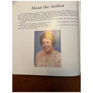 May include: A page from a book titled "About the Author" features a portrait of a smiling woman with short, dark hair. The text describes her career as a writer and Barbie doll collector. The page is open on a wooden surface.