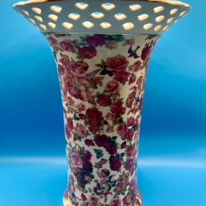 May include: A cream-colored ceramic vase featuring a vibrant floral pattern of pink and red roses interspersed with small winged cherubs. The vase has a wide, flared top rim with a decorative lattice cutout design and a thin gold-tone trim.