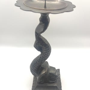 May include: A dark bronze-colored candle holder with a floral-shaped top and a curved, textured stem. The base is square with decorative detailing. The candle holder is designed to hold a single candle.