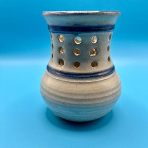 May include: A ceramic vessel with a flared rim and a rounded base, featuring a speckled off-white glaze. The upper section is adorned with three horizontal rows of circular cutouts and two distinct dark blue painted bands encircling the body.