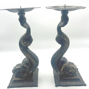 May include: Pair of antique bronze candle holders. Each holder features a stylized serpent design with a floral-shaped candle cup. The base is a rectangular shape with a decorative edge. The candle holders are a dark bronze color.
