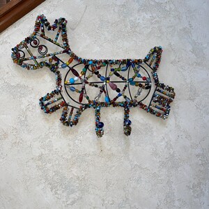 May include: A decorative dog-shaped wall hanging made from a metal frame and colourful beads. The beads are in various colours including blue, green, red, and yellow. The dog has a playful design with a tail, legs, and a head with curly details.