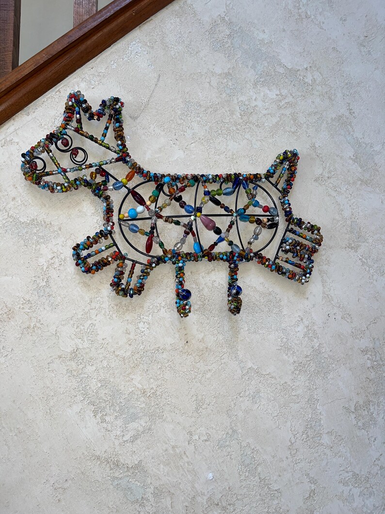 May include: A decorative dog-shaped wall hanging made from a metal frame and colourful beads. The beads are in various colours including blue, green, red, and yellow. The dog has a playful design with a tail, legs, and a head with curly details.
