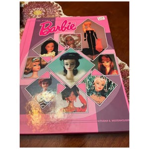 May include: A pink book titled "The Story of Barbie" with the Barbie logo in cursive. The cover features images of Barbie dolls in various styles and eras. The book is a vintage collectible.