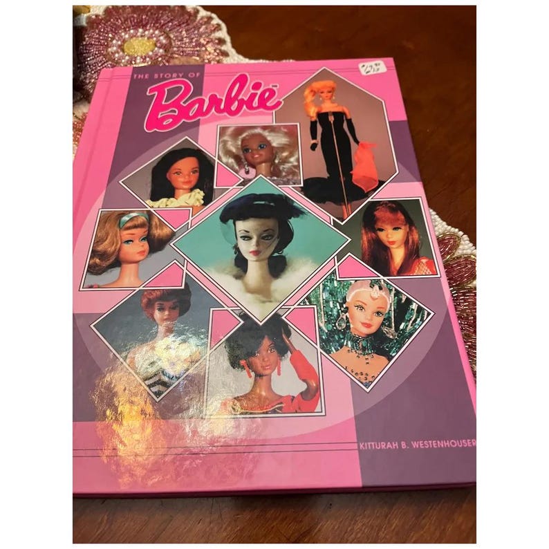 May include: A pink book titled "The Story of Barbie" with the Barbie logo in cursive. The cover features images of Barbie dolls in various styles and eras. The book is a vintage collectible.