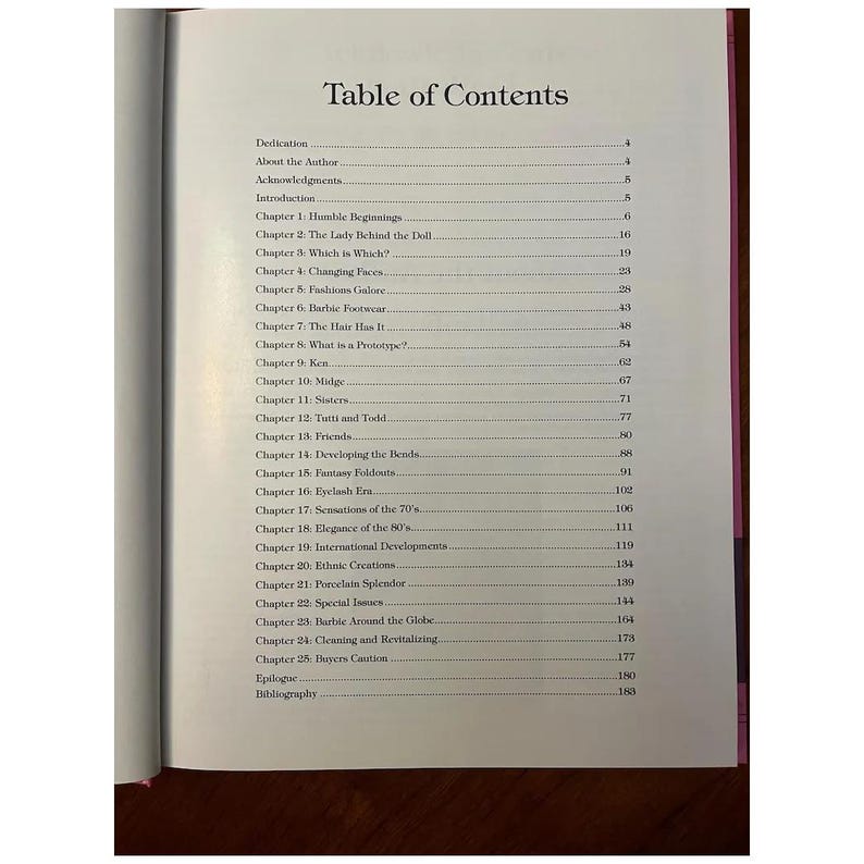 May include: Open book page with a table of contents titled "Table of Contents." The list includes chapter titles such as "Humble Beginnings," "Changing Faces," and "Barbie Footwear." The book has a pink spine.