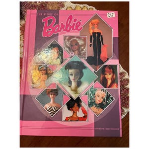 May include: A pink book titled "The Story of Barbie" with a collage of Barbie doll images. The cover features the Barbie logo in pink and a price tag in the upper right corner. The book's cover is framed with pink borders.