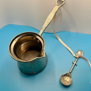 May include: A silver-colored Turkish coffee pot with an ornate handle and a small decorative spoon. The pot has a wide opening and a spout for pouring. The spoon has a floral design on the bowl and a decorative handle. A light pink ribbon is attached to both items.