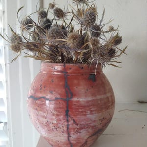 May include: A round, red ceramic vase with a speckled pattern and dark gray lines, holding dried thistle flowers. The vase has a wide opening and a bulbous shape, sitting on a white surface.