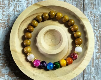 May include: A beaded bracelet with a mix of wooden and colorful gemstone beads. The bracelet is displayed on a small, round wooden dish. The wooden beads are a warm brown color, and the gemstone beads are various colors.