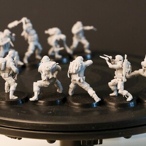 May include: A group of small, grey soldier figurines in dynamic poses, each on a black base. They are displayed on a black rotating stand. The background is dark, highlighting the figures' details.