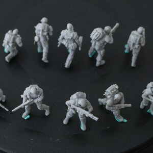 May include: Ten miniature grey soldier figurines, each with backpacks and rifles, are displayed. The figures are in various action poses, with small green accents on their feet. They are arranged on a dark, circular surface.