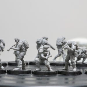 May include: A collection of gray miniature figurines depicting soldiers in combat gear. Each figure is mounted on a black base and carries a weapon or backpack. The figures are detailed and appear to be made of plastic.