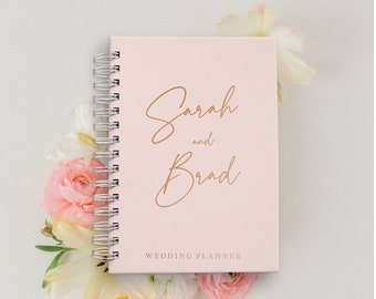 May include: A pink wedding planner with a gold inscription that reads "Sarah and Brad" and the words "Wedding Planner" printed below.