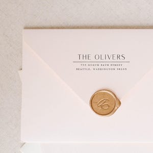 May include: A pale pink envelope featuring black printed text that reads THE OLIVERS, 753 NORTH 84TH STREET, SEATTLE, WASHINGTON 98103. The envelope is sealed with a circular gold wax seal embossed with a cursive letter C in the center.