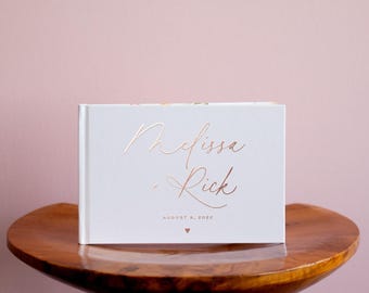 May include: A white landscape-oriented guest book featuring rose gold foil lettering that reads Melissa + Rick, with the date August 9, 2022, and a small heart symbol printed below the text in a matching rose gold metallic finish.