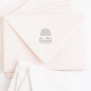 May include: A stack of pale pink envelopes with a black ink stamp on the top envelope. The stamp features a line-art rainbow arch above the text: claire + thomas, 755 NORTH 84TH STREET, SEATTLE, WASHINGTON, 98103, printed in a serif font.