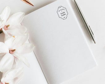 May include: An open white notebook featuring a black ink stamp with an ornate border and the text From the Library of Emilie Nadler. The notebook is accompanied by a metallic silver pen, a rose gold pen, rose gold glasses, and white flowers.