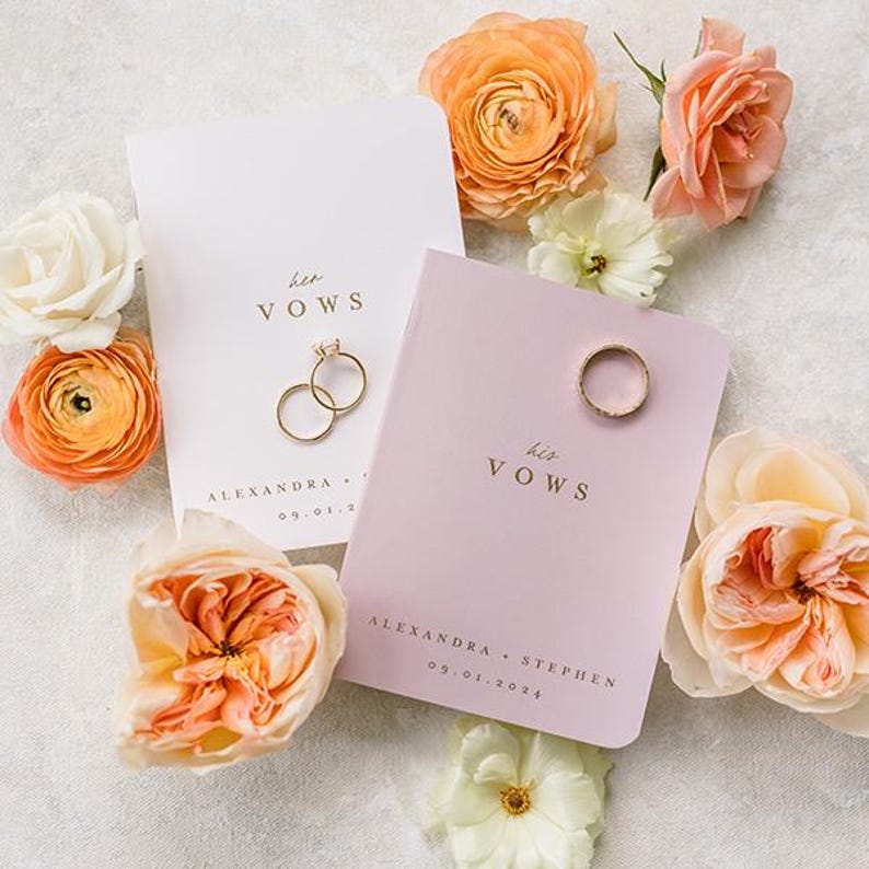 May include: Two vow booklets, one white and one light purple, featuring gold text that reads her VOWS and his VOWS, along with the names Alexandra and Stephen and the date 09.01.2024. Two gold rings rest on the booklets, surrounded by peach roses.