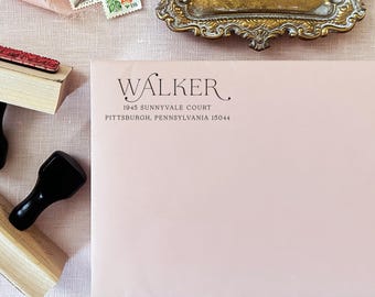 May include: A pale pink envelope featuring black printed text that reads WALKER, 1945 SUNNYVALE COURT, PITTSBURGH, PENNSYLVANIA 15044. The envelope is positioned next to two wooden rubber stamps and a small ornate gold metal tray.