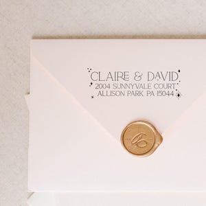 May include: A pale pink envelope featuring black printed text that reads CLAIRE & DAVID 2004 SUNNYVALE COURT ALLISON PARK PA 15044 with small star icons. The envelope is sealed with a round gold wax seal featuring an embossed cursive letter design.