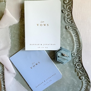 May include: A white booklet with gold text reading her VOWS and a light blue booklet with black text reading his VOWS, both personalized with HANNAH & JONATHAN 06.10.23. A hexagonal blue velvet ring box holds two silver rings with stones.