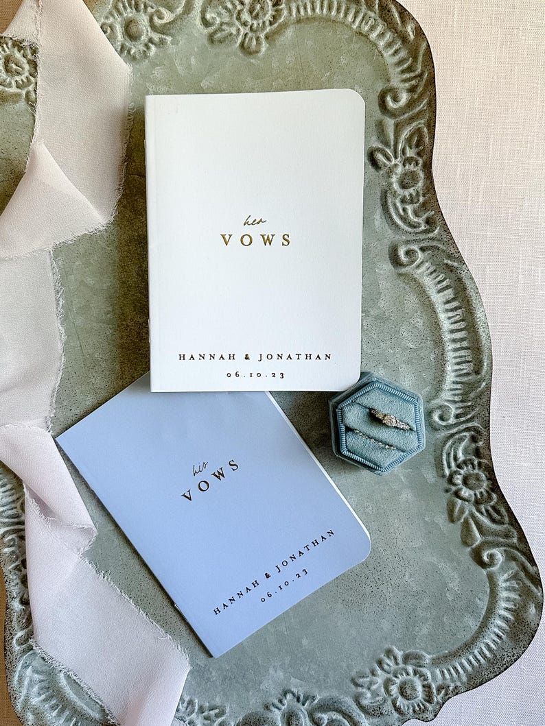 May include: A white booklet with gold text reading her VOWS and a light blue booklet with black text reading his VOWS, both personalized with HANNAH & JONATHAN 06.10.23. A hexagonal blue velvet ring box holds two silver rings with stones.