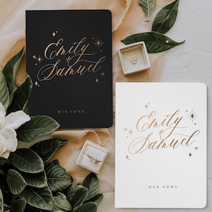 May include: Two black and white vow books with gold lettering that reads "Emily + Samuel" and "His Vows" and "Her Vows". The books are surrounded by greenery and a ring box.