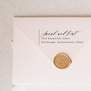 May include: A pale pink envelope featuring a black printed return address that reads Sarah and Pat, 1945 Sunnyvale Court, Pittsburgh, Pennsylvania 15044. The envelope is sealed with a round, metallic gold wax seal featuring an embossed script letter.